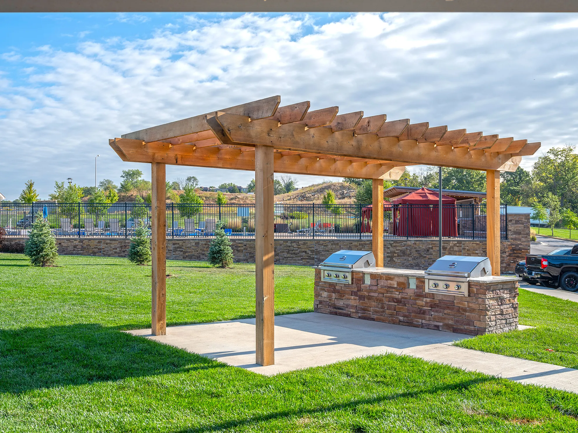 Inviting outdoor space featuring a modern pergola with built-in stone grills, surrounded by lush green lawn and vibrant landscape.