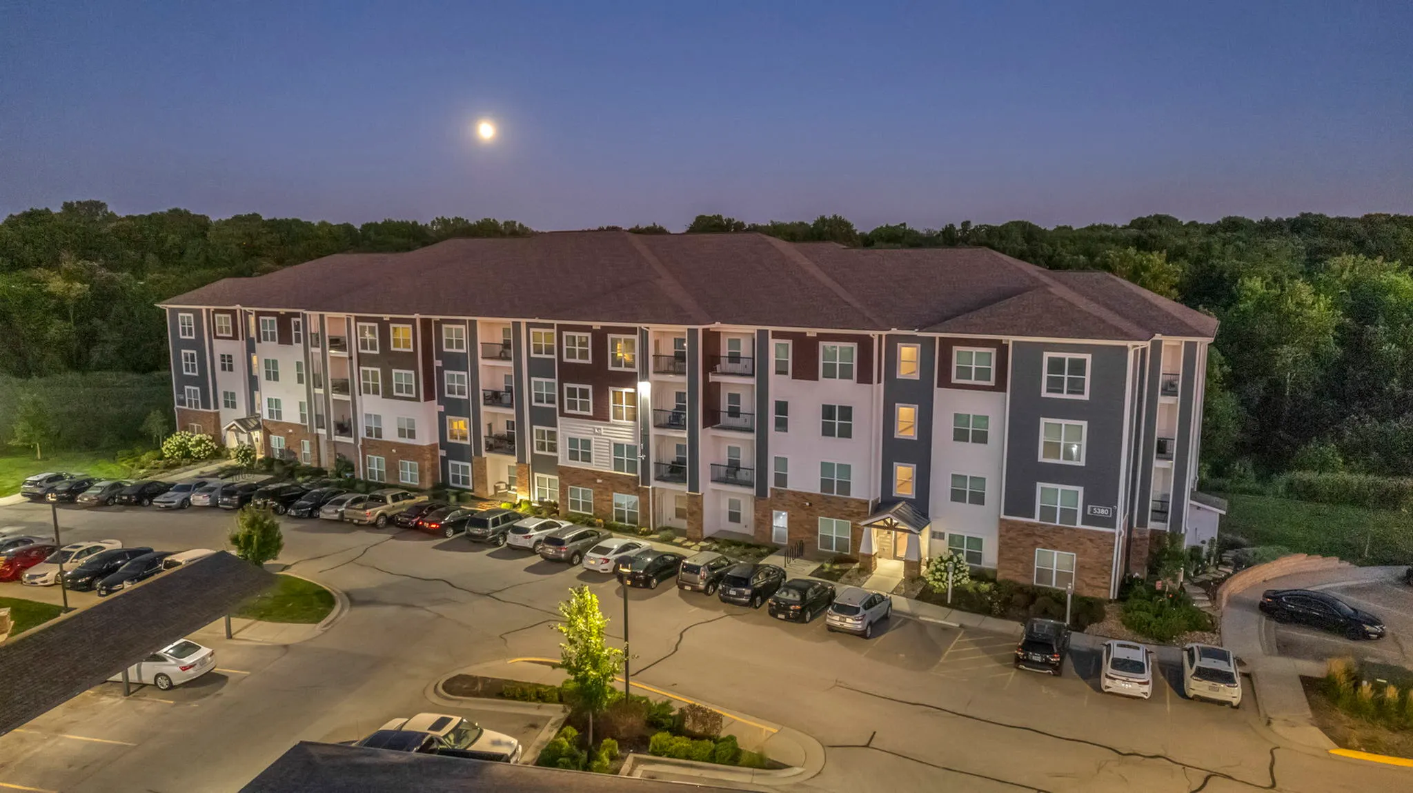 Elegant apartment building beautifully illuminated under a serene night sky, surrounded by lush greenery and featuring ample parking.