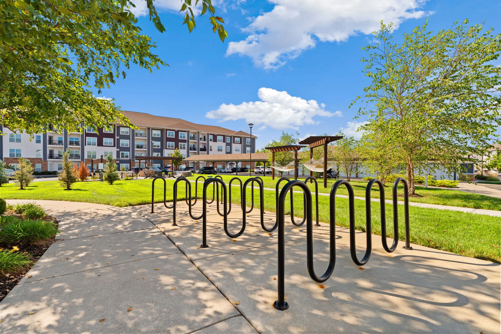 Inviting apartment community with well-maintained gardens, modern bike racks, and relaxing hammock area under a bright blue sky.