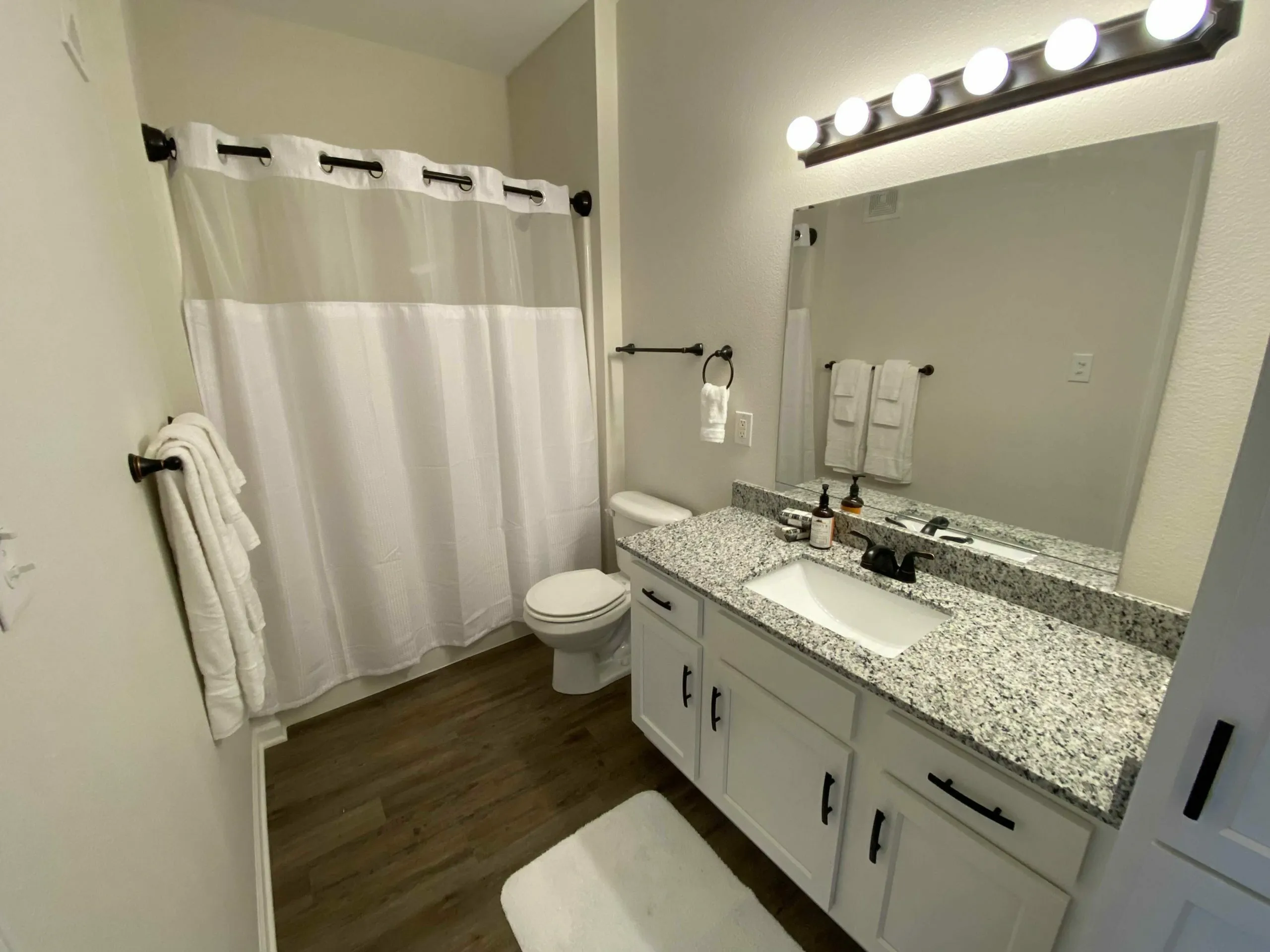 Modern bathroom with elegant granite countertop, stylish white cabinetry, and warm wood flooring, featuring a sleek lighted mirror and a crisp white shower curtain for a luxurious touch.