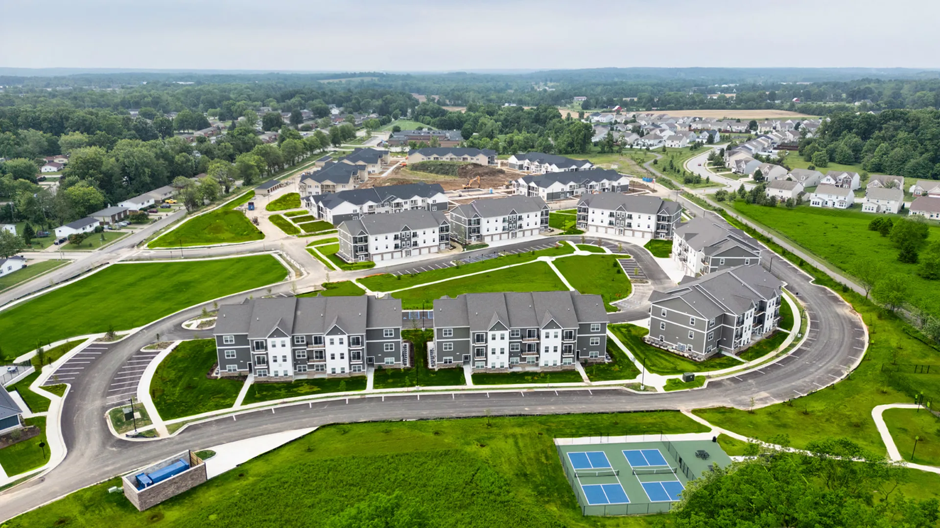 Aerial view of modern apartment complex with well-manicured lawns, surrounded by lush greenery, featuring ample parking and tennis courts for an active lifestyle.