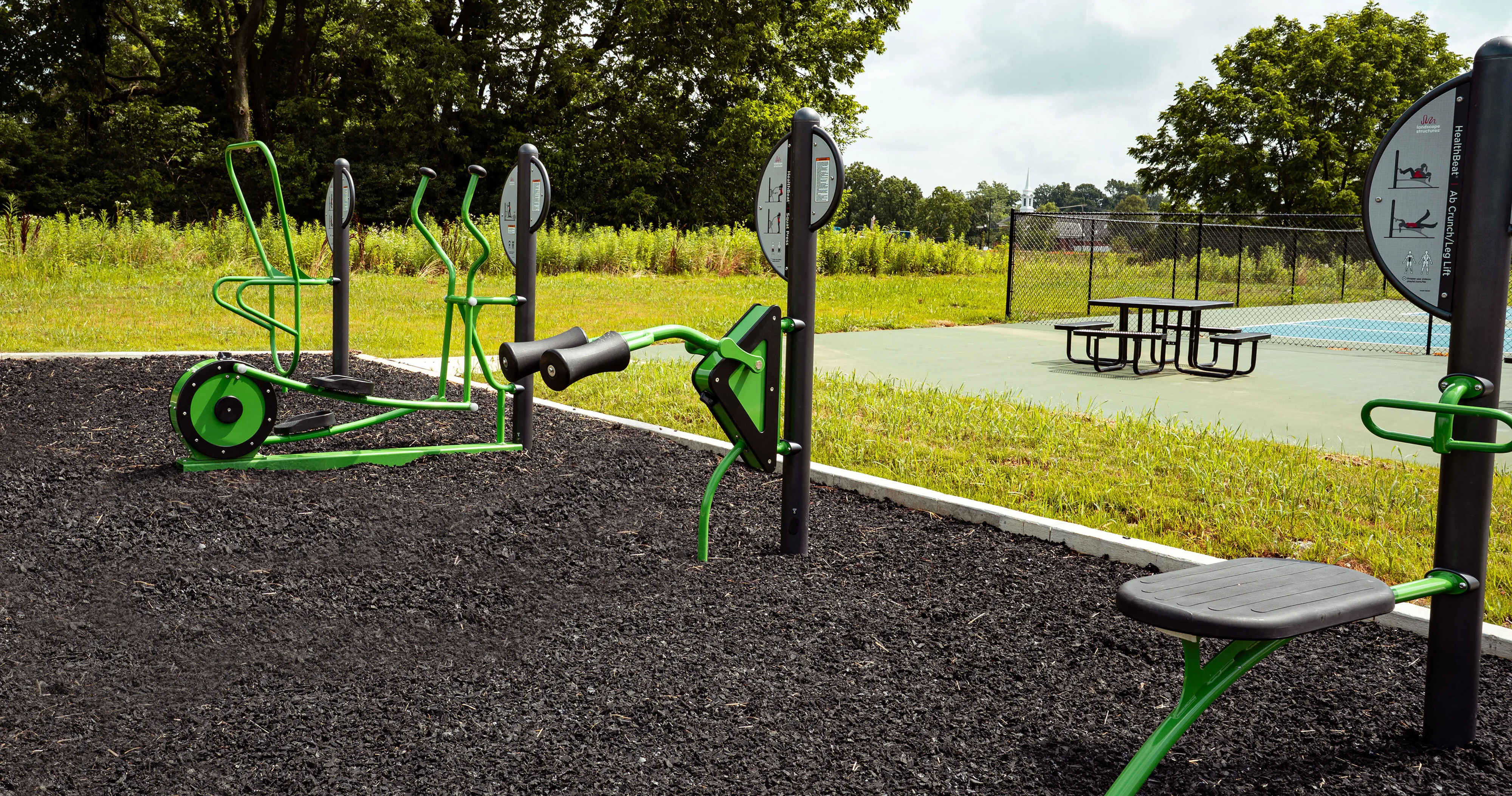 Inviting outdoor fitness area featuring modern green exercise equipment and a picnic table, set against a lush natural backdrop.