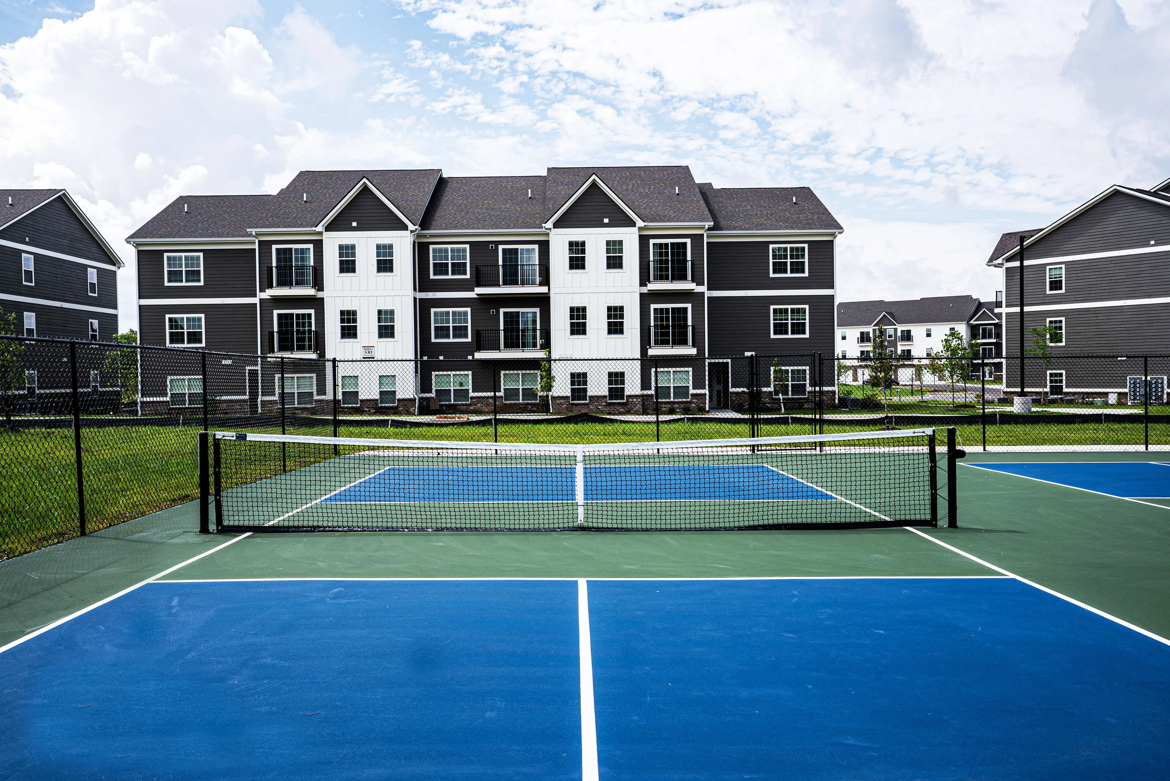 Modern apartment complex with elegant design, featuring a pristine tennis court and lush green surroundings.