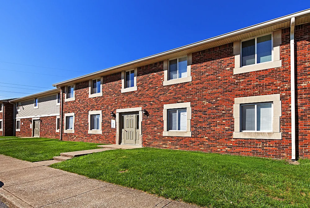 Charming two-story brick apartment building with neatly landscaped entrance under a clear blue sky.