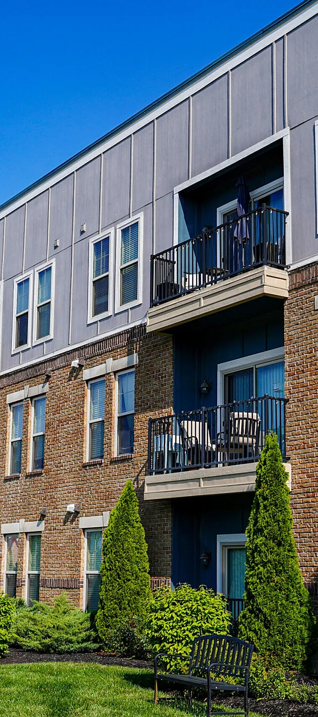 Modern apartment building with brick facade, balconies, and lush greenery on a sunny day.