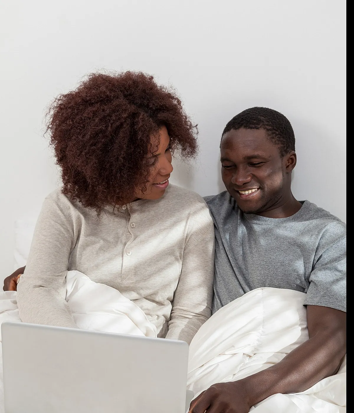 Cozy couple enjoying a relaxed morning in bed with a laptop, surrounded by plush white bedding.