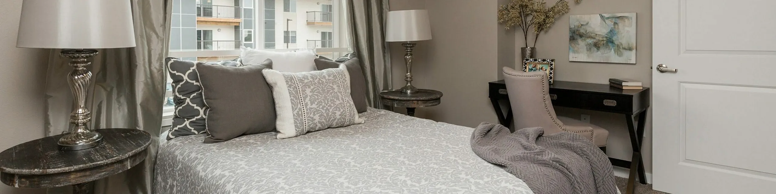 Charming bedroom with elegant gray patterned bedding, natural light, and a stylish desk space in a modern apartment.