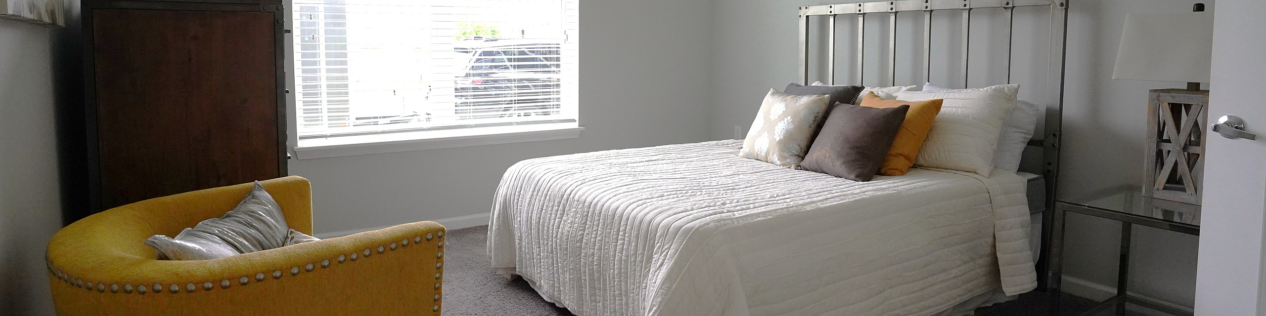 Bright and modern bedroom featuring a comfortable bed with soft, layered pillows, charming yellow accent chair, and large window that lets in natural light.