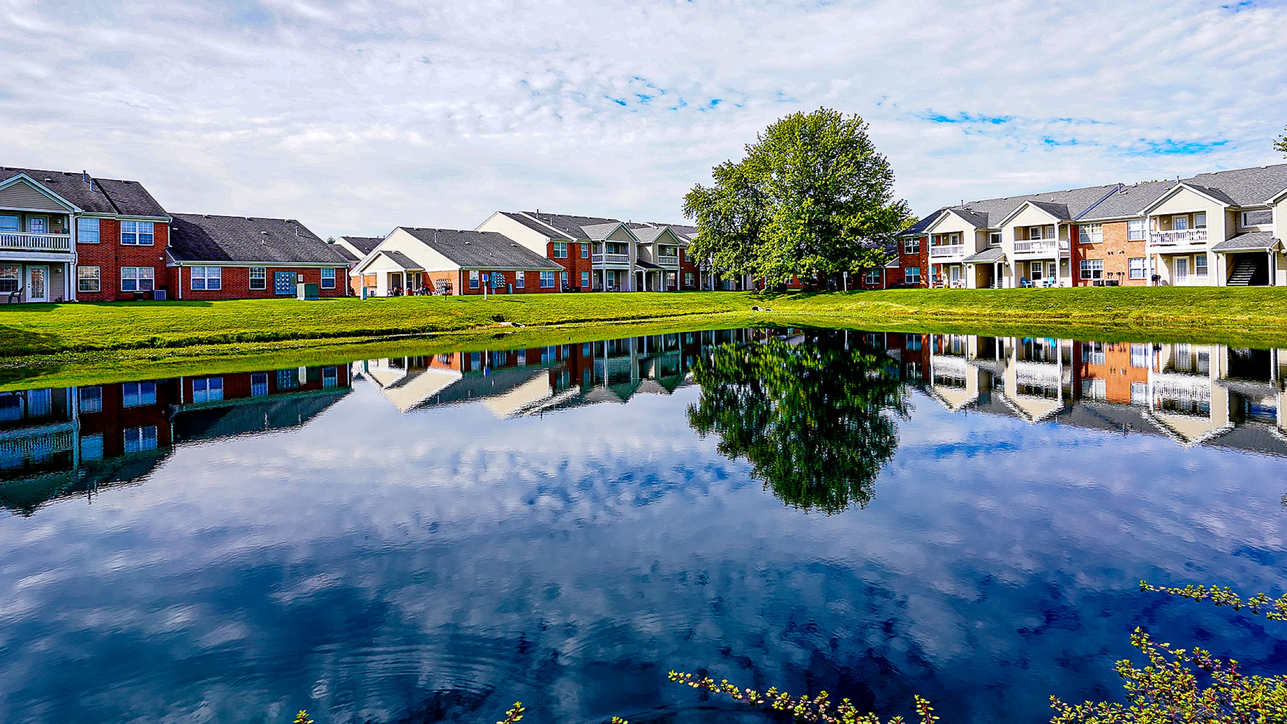 Idyllic apartment complex with lush green lawns, serene pond reflections, and modern architecture under a vibrant sky.