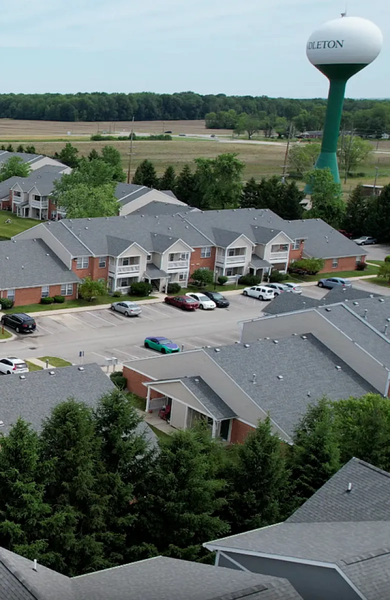 Scenic aerial view of charming residential community with modern apartments, lush greenery, and a tranquil pond, perfect for serene living.