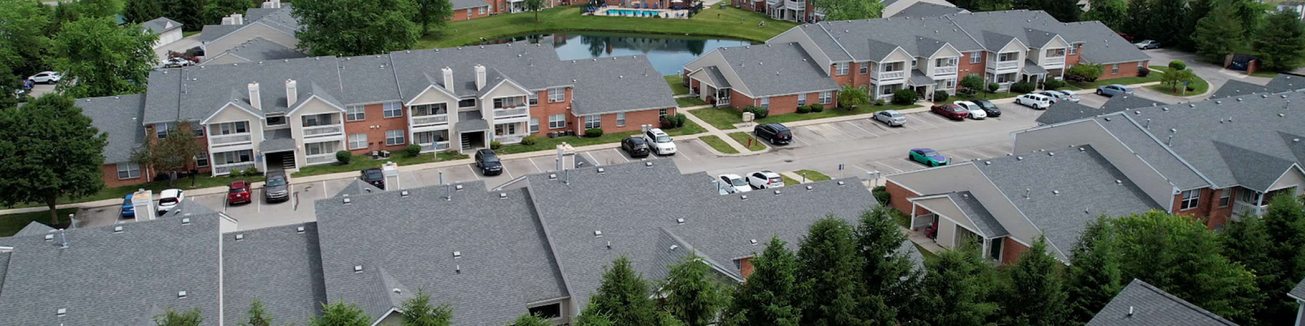 Charming community aerial view featuring modern apartments, lush greenery, and a serene pond, offering a peaceful suburban lifestyle.