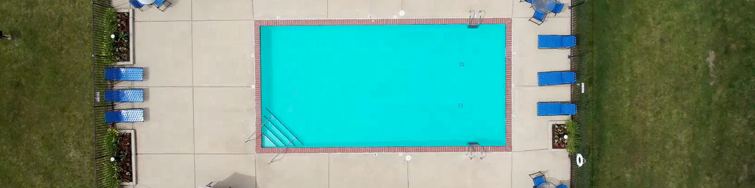 Aerial view of a vibrant outdoor swimming pool surrounded by lush greenery, featuring blue loungers and patio tables for relaxation and leisure.