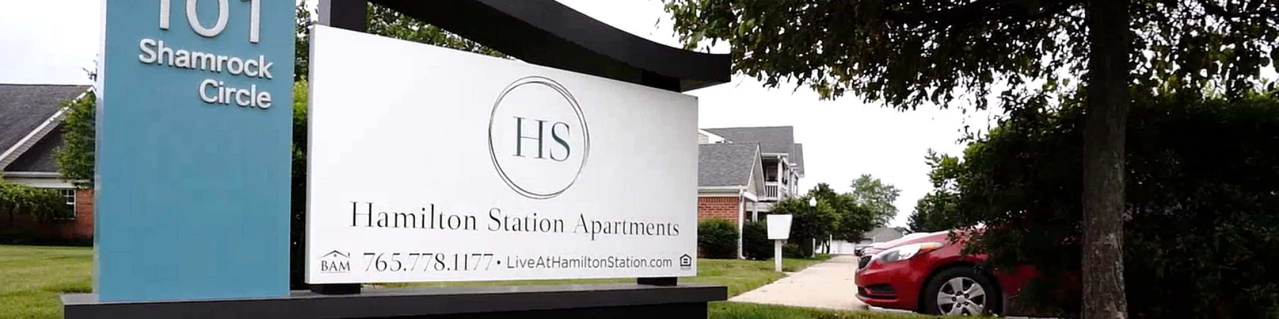 Inviting entrance sign for Hamilton Station Apartments at 101 Shamrock Circle, surrounded by vibrant flowers and lush greenery.