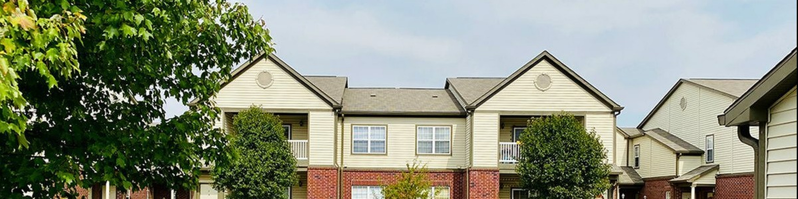 Charming two-story brick and siding apartment complex with balconies, surrounded by lush greenery and a serene atmosphere.