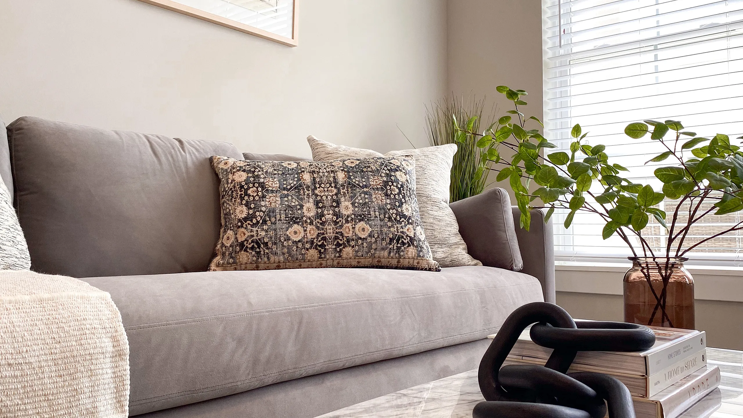 Elegant living room with a plush gray sofa, stylish patterned cushions, and a marble coffee table adorned with chic decor. Sunlit room with vibrant green plant and modern artwork creating a cozy, inviting space.