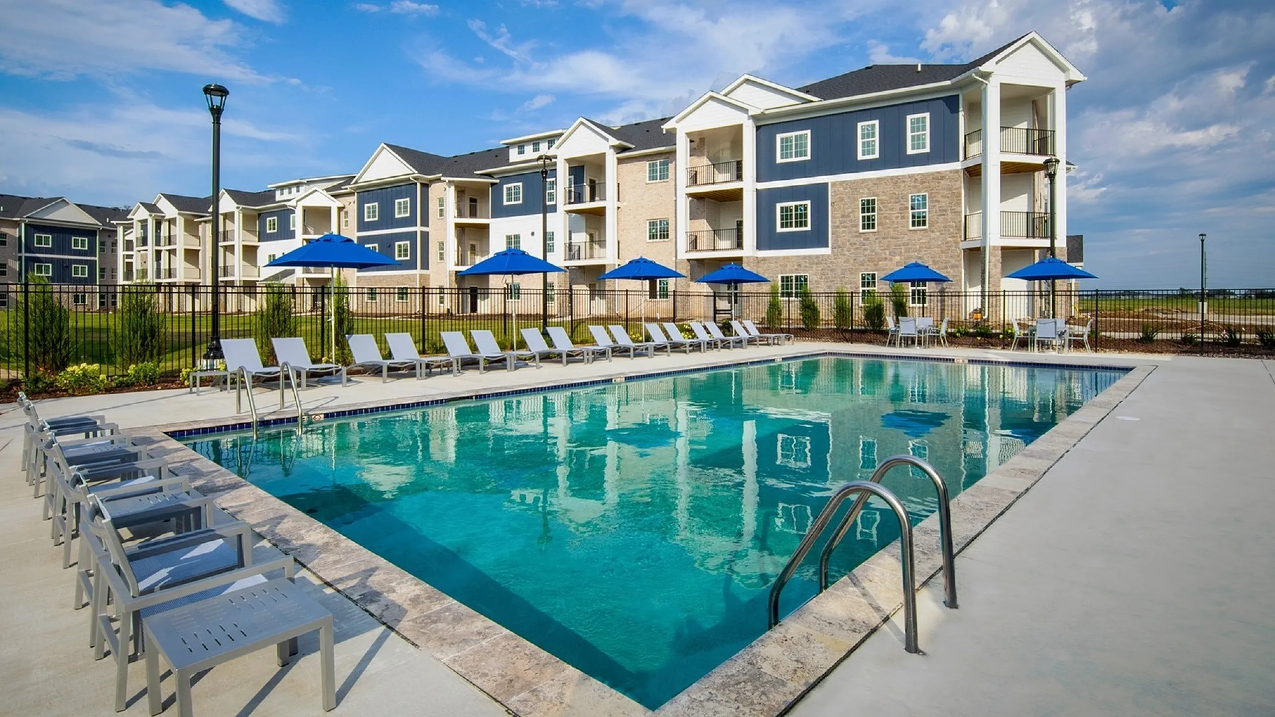 Luxurious apartment complex featuring a pristine outdoor pool with comfortable seating and shaded blue umbrellas, set against a backdrop of modern architecture and clear blue skies.