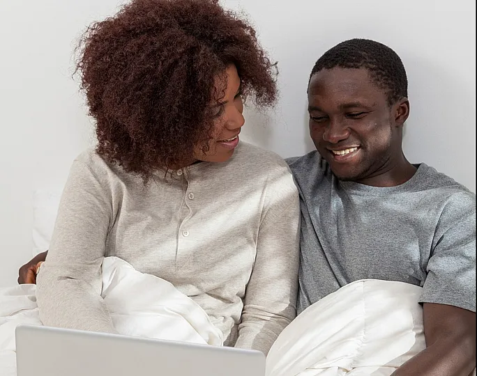 Cozy couple enjoying a relaxed moment on a bed, browsing their laptop together in a bright, white bedroom.