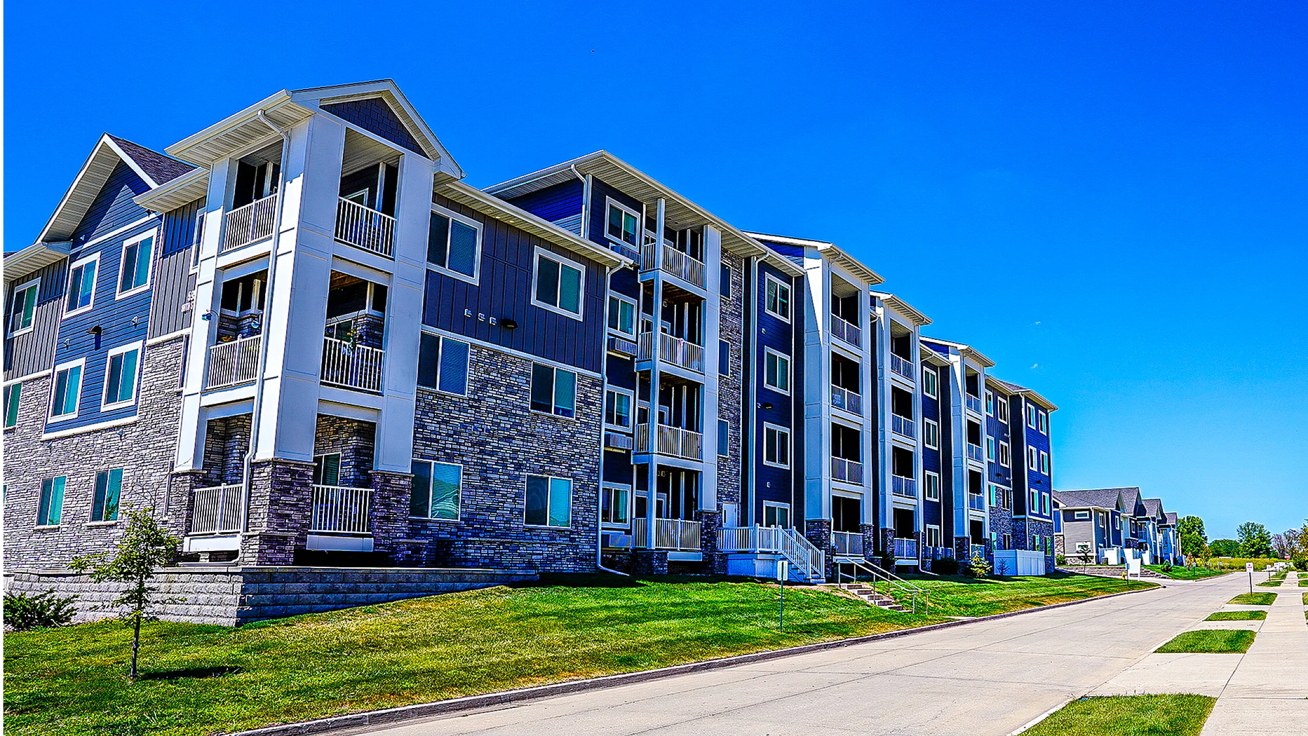 Modern apartment complex with stylish stone facade and spacious balconies, set against a clear blue sky. Ideal living environment with well-maintained green lawns and inviting walkways.