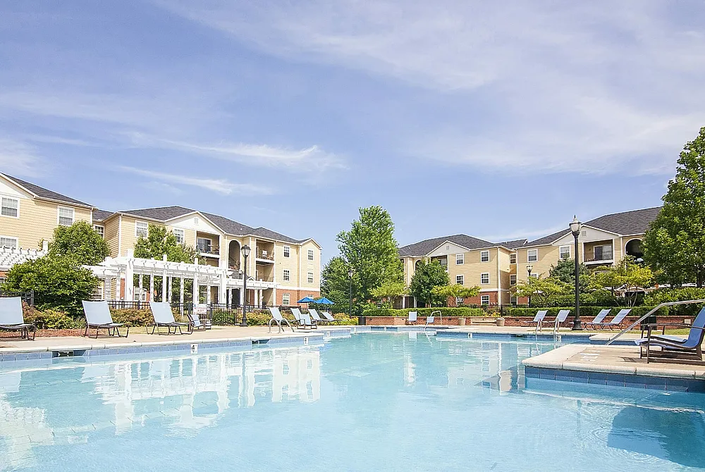 Vibrant apartment complex featuring a spacious pool, surrounded by elegant lounge chairs and lush greenery under a clear blue sky.