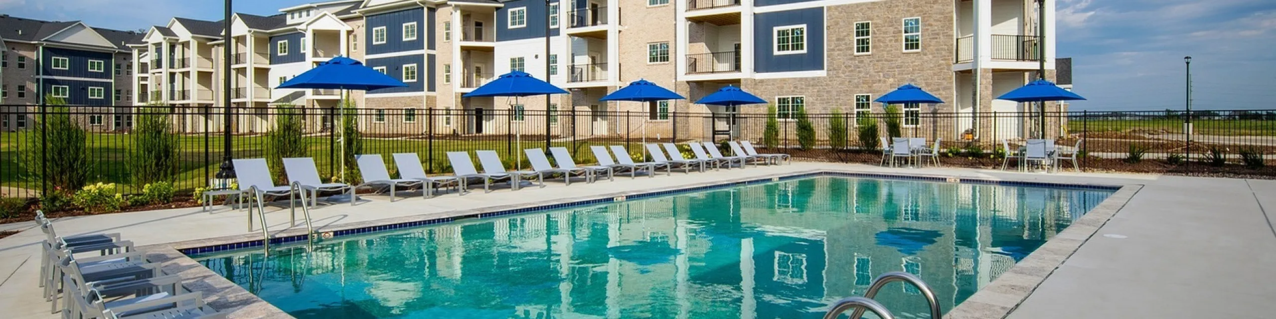 Luxurious apartment complex with a pristine swimming pool, surrounded by modern buildings and lush greenery, under a clear blue sky.
