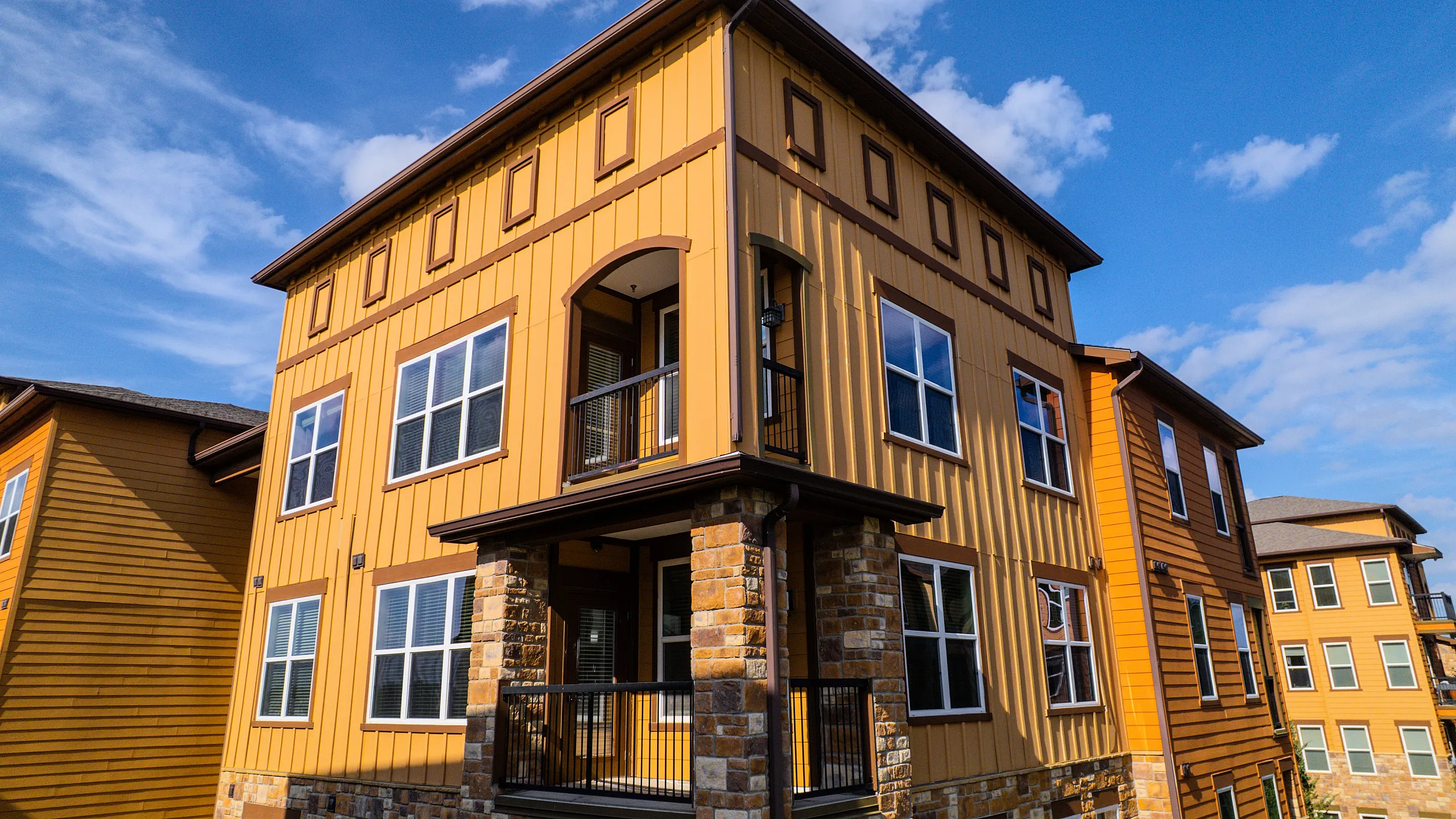 Modern apartment building with yellow and brown siding, featuring large windows and a welcoming balcony under a bright blue sky.