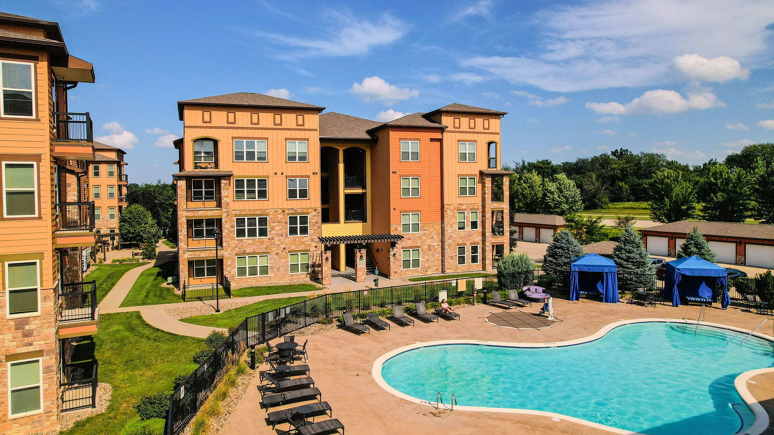 Stunning apartment complex with modern architecture, lush greenery, and a sparkling pool surrounded by lounge chairs and blue cabanas.