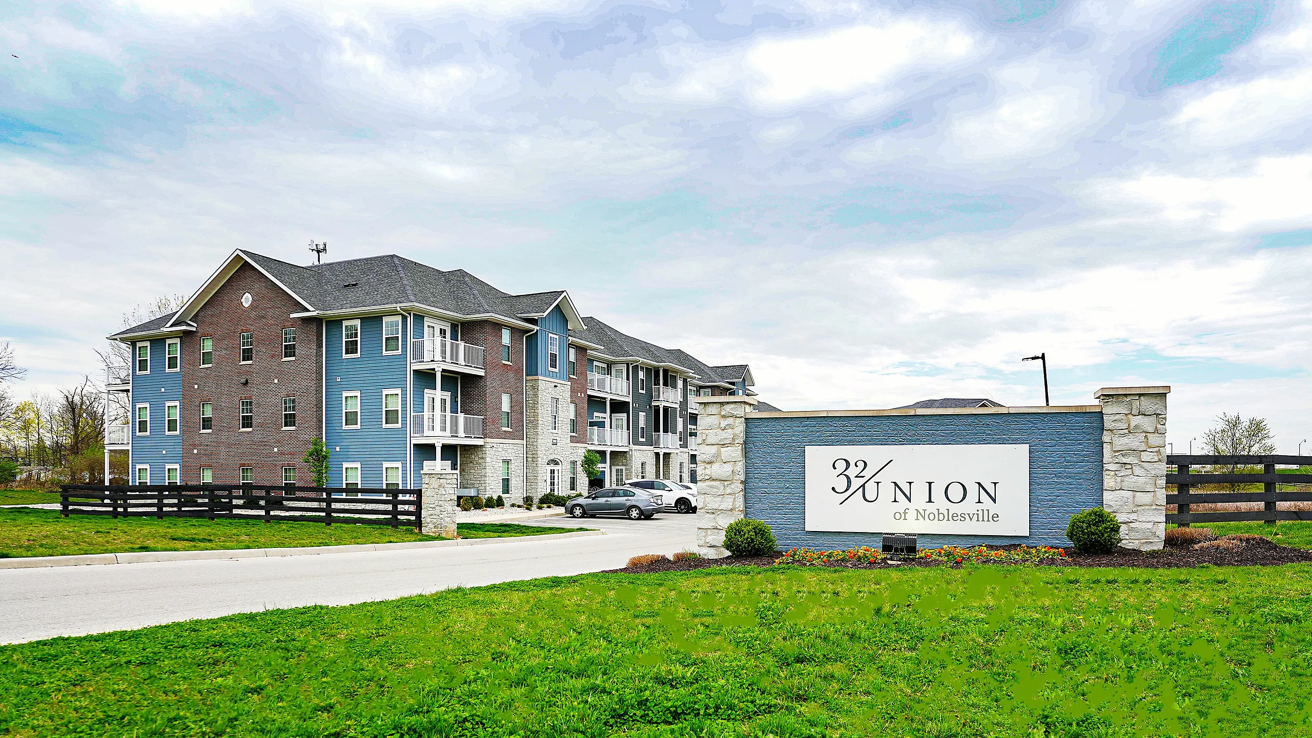 Elegant apartment complex at 32 Union of Noblesville featuring modern design with blue and brick exterior, surrounded by lush green landscape.