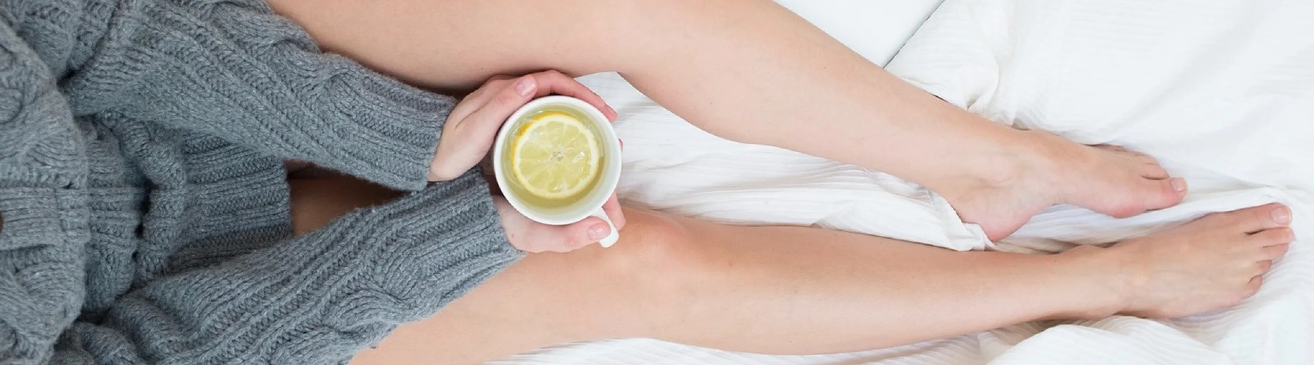 Cozy scene of a person relaxing on a bed with a cup of lemon tea, wrapped in a warm grey sweater, next to a sleek laptop.