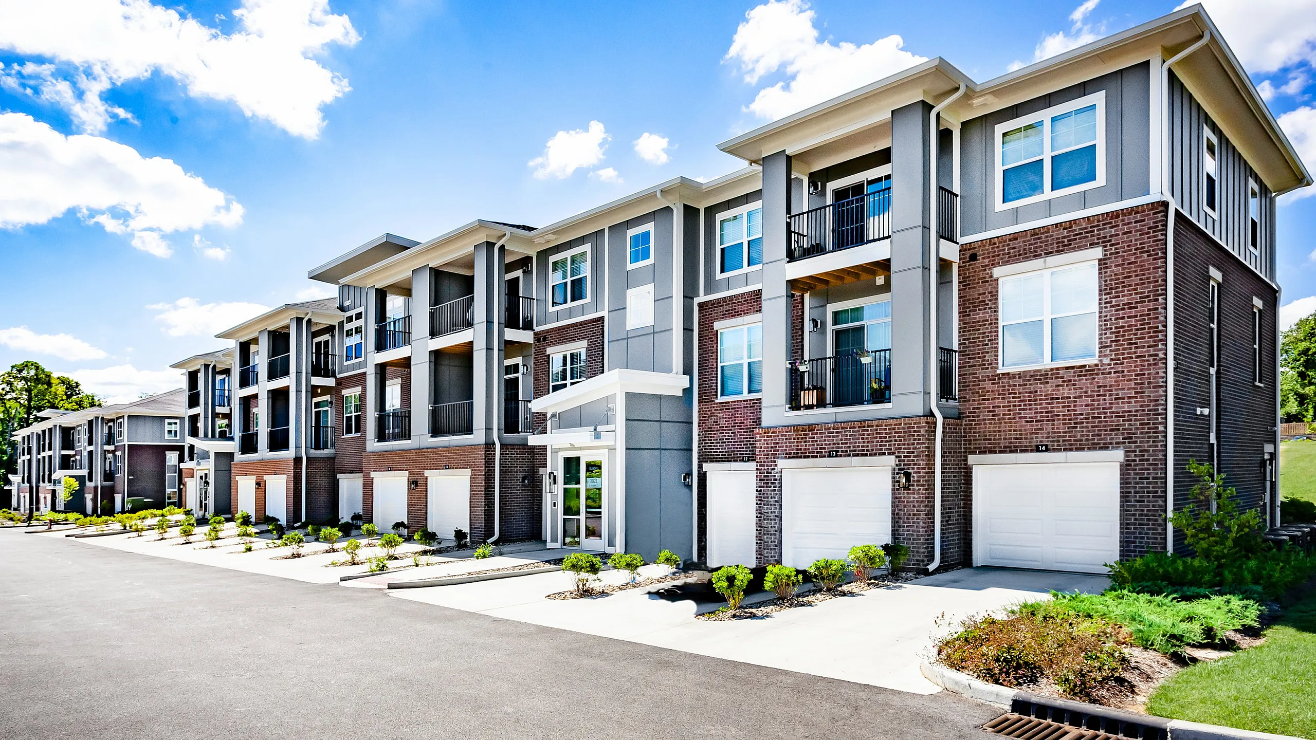 Modern apartment complex with a stylish brick facade, featuring balconies and lush landscaping, set against a bright blue sky.