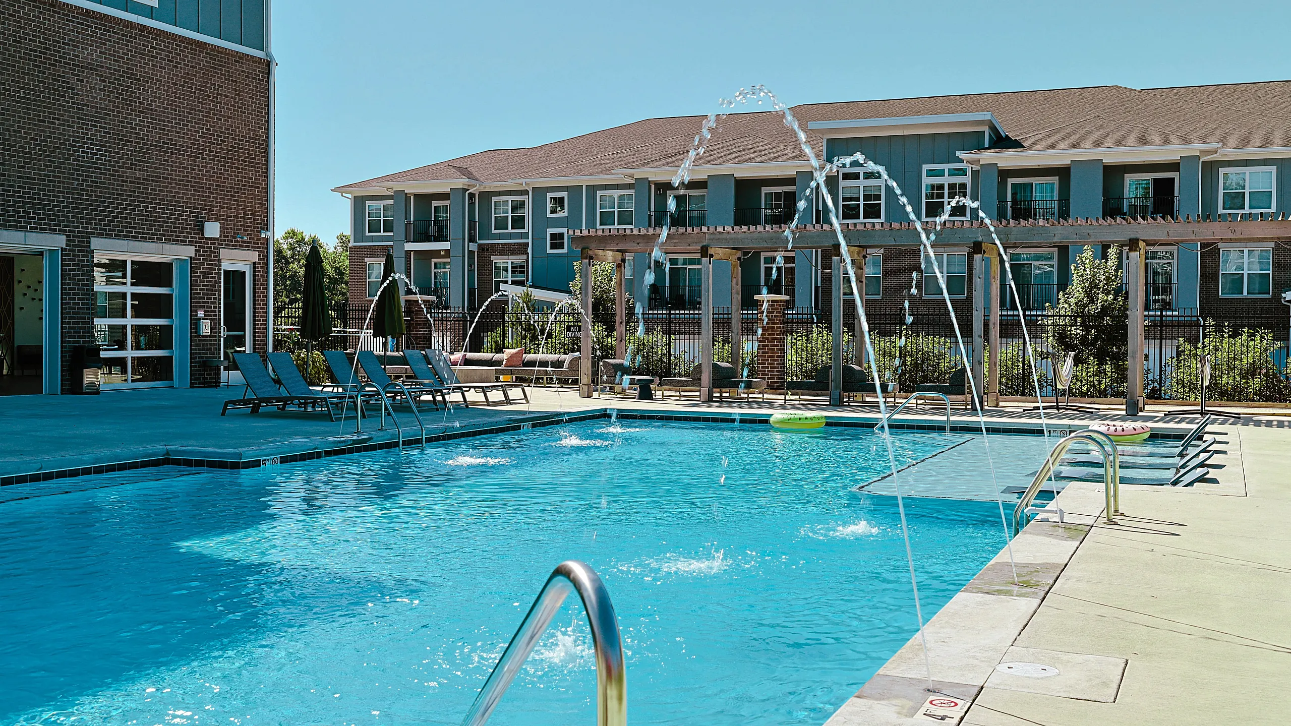 Luxurious apartment complex featuring a sparkling swimming pool with water fountains, surrounded by comfortable lounge chairs and modern buildings.