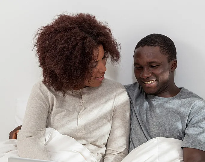 Cozy bedroom scene with a couple happily using a laptop under soft, white bedding in a bright, minimalist setting.