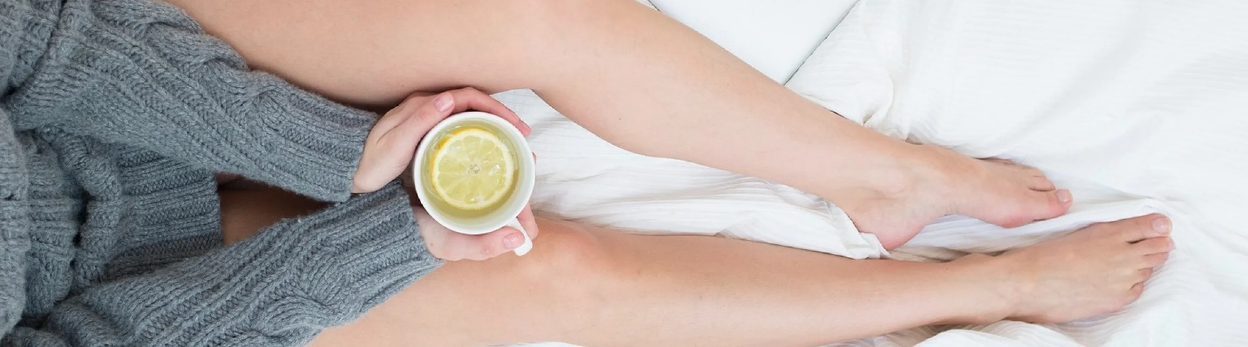 Cozy and elegant setting with a person relaxing in a soft, white bed, holding a cup of tea with a lemon slice, beside a sleek silver laptop.
