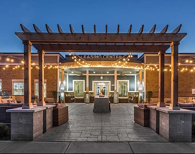 Charming leasing center entrance with warm string lights and cozy outdoor seating under a wooden pergola at dusk.