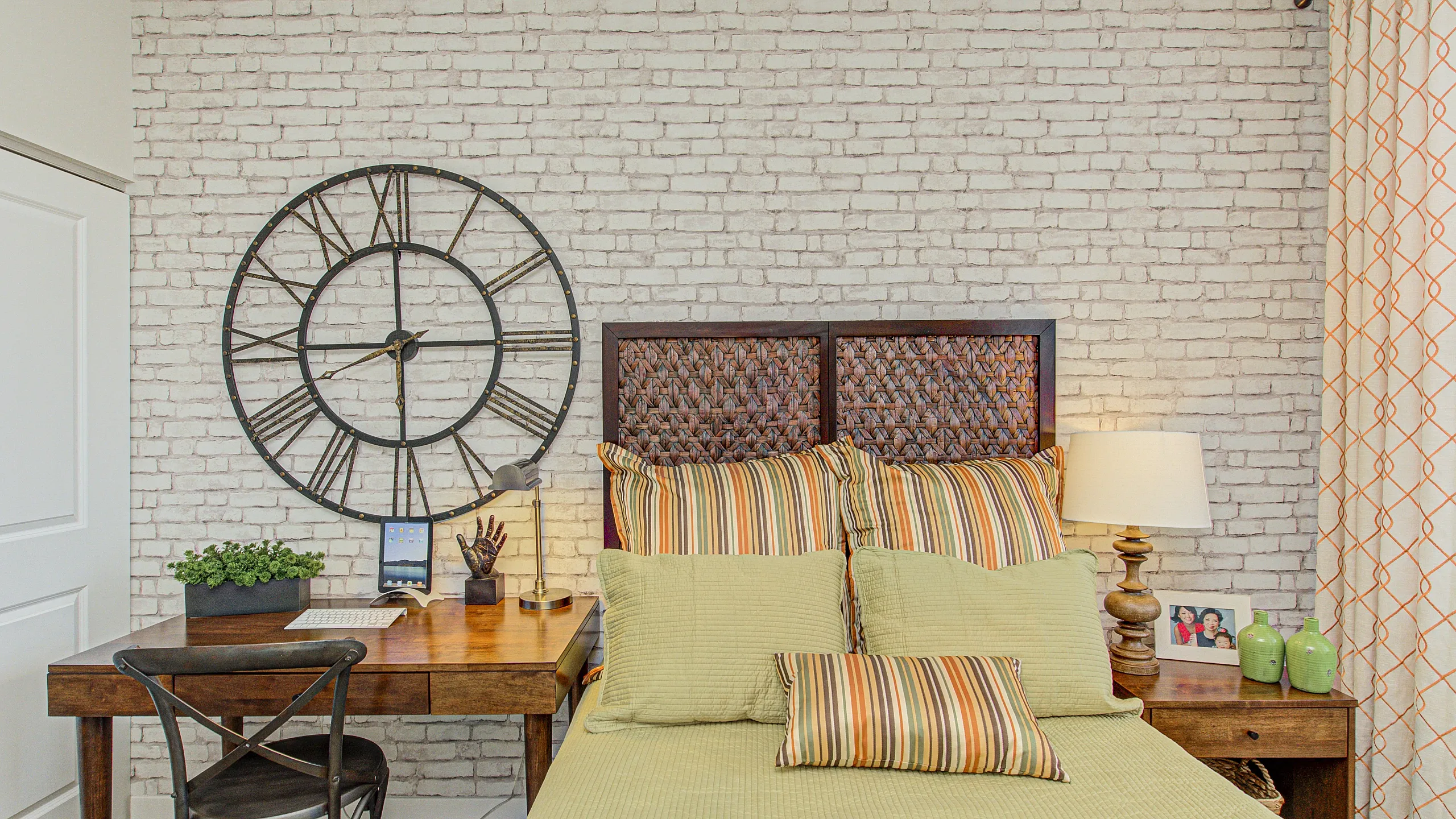 Cozy bedroom with a stylish exposed brick wall, featuring a vintage-style oversized clock, elegant wooden desk, and a comfortable bed with striped pillows for a charming, inviting atmosphere.