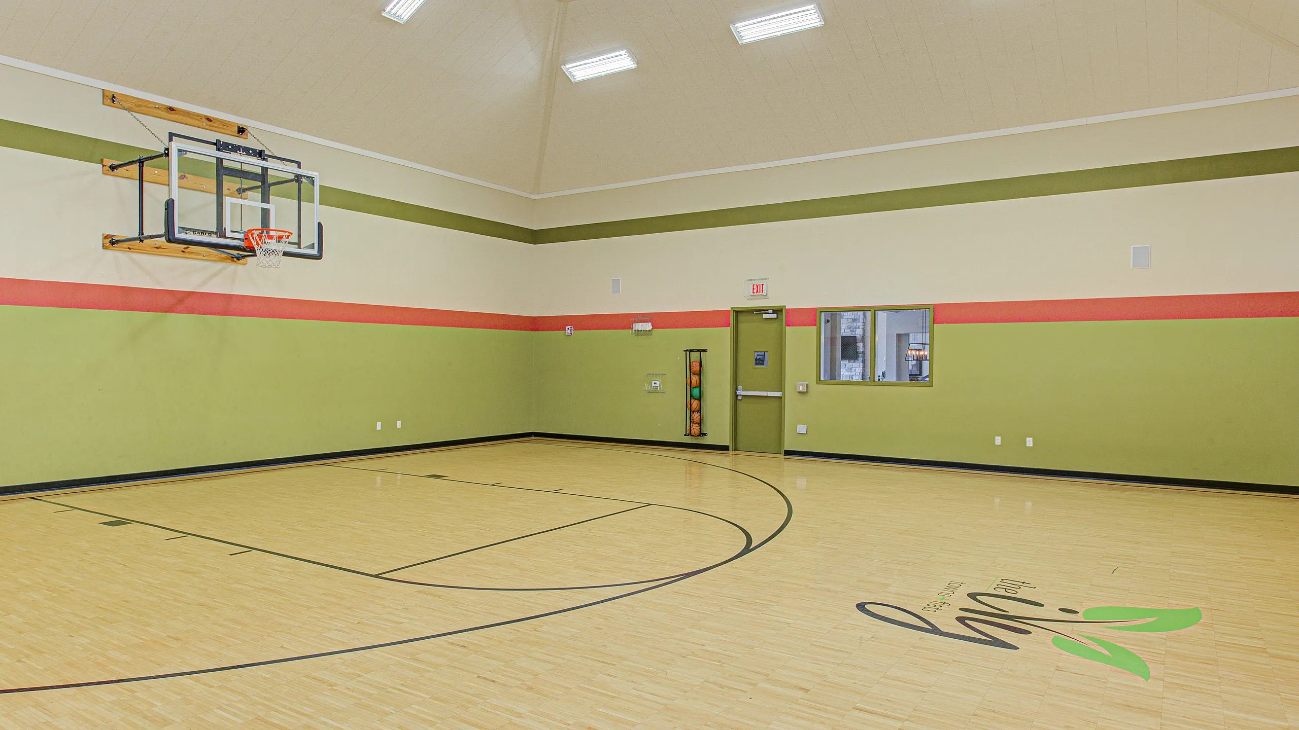 Modern indoor basketball court with polished wooden floors, vibrant green and red walls, and ample overhead lighting.