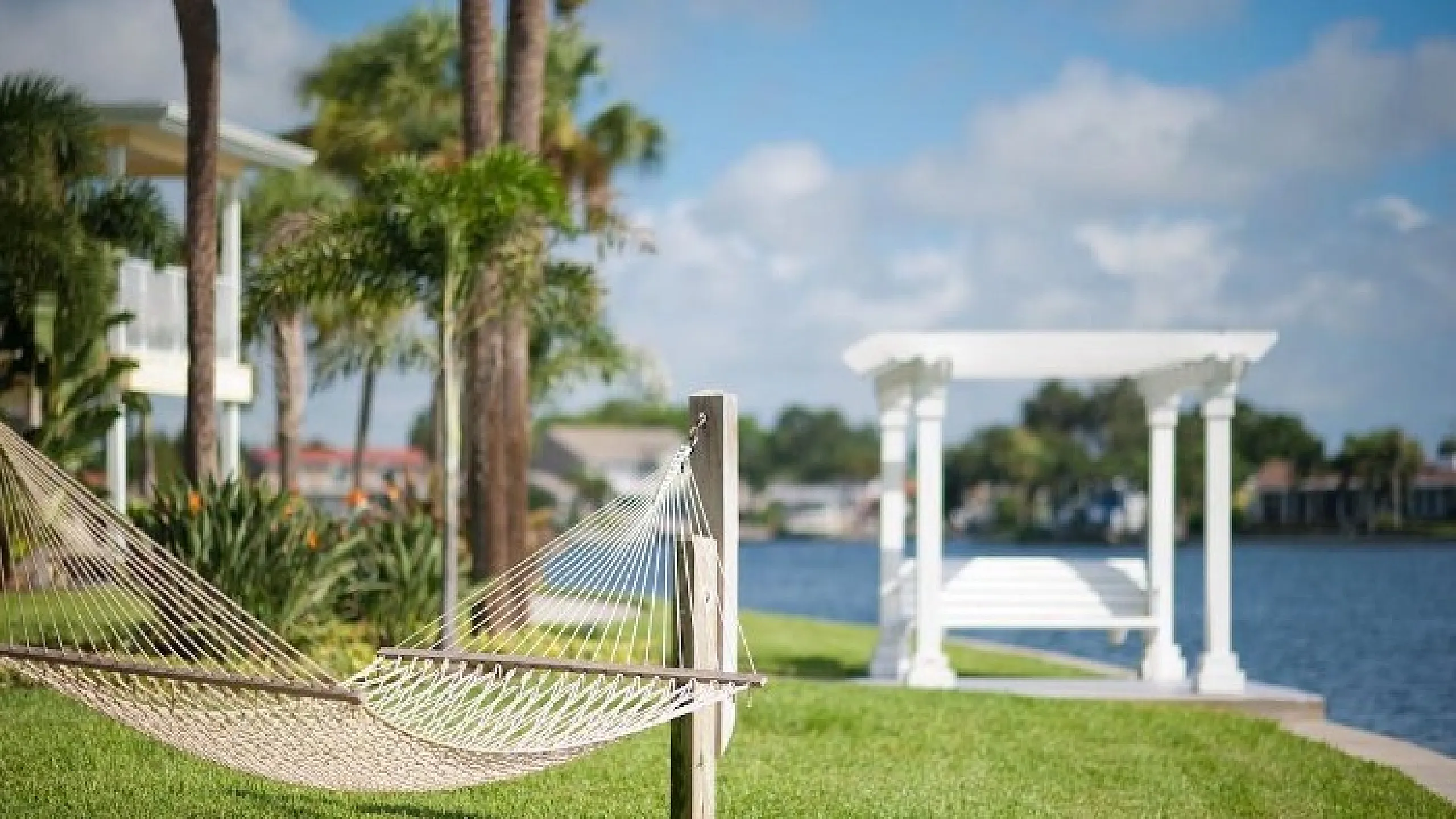 Peaceful lakeside hammock with lush greenery and a white pergola under a vibrant sky.