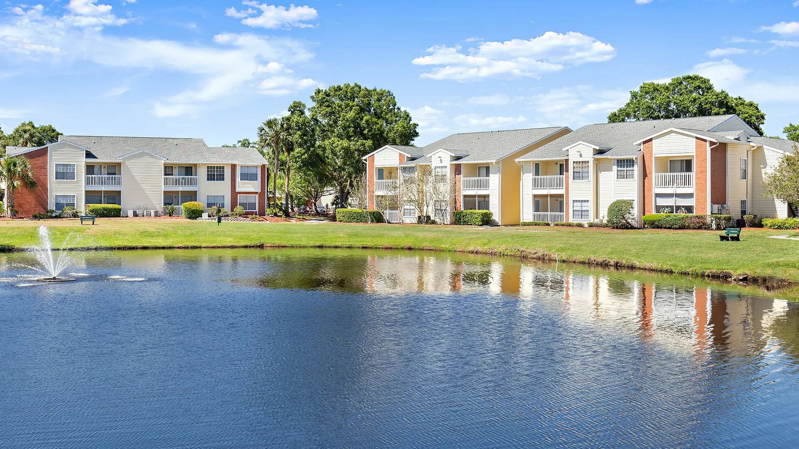 Charming lakeside apartment complex with manicured lawns and a sparkling water fountain in the foreground.
