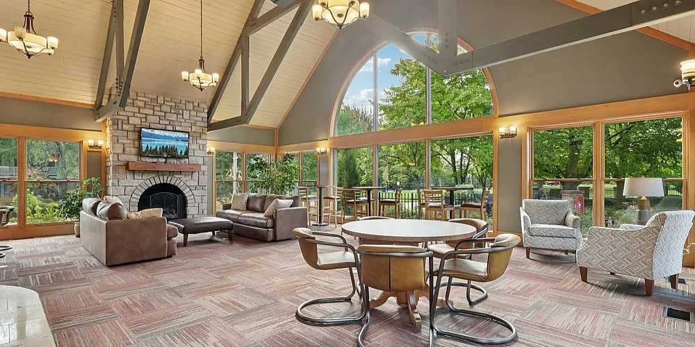 Inviting communal lounge with large windows, cozy seating, and a stunning stone fireplace overlooking lush greenery.