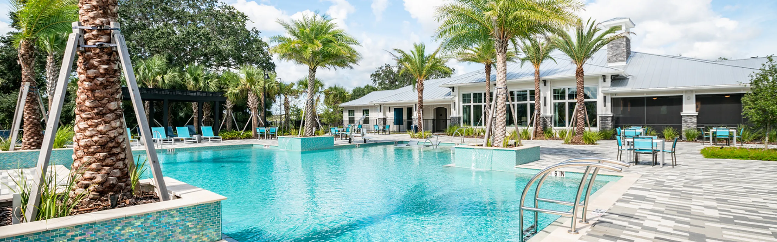 Luxurious apartment community with a sparkling pool, surrounded by palm trees and modern outdoor seating, under a bright blue sky.