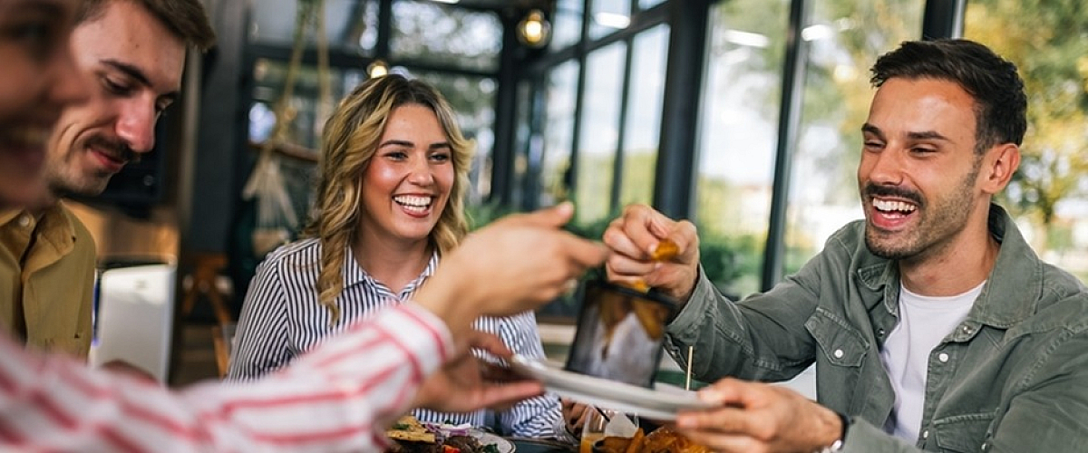Joyful group of friends enjoying a meal together in a bright, modern dining area with large windows and lush greenery outside.