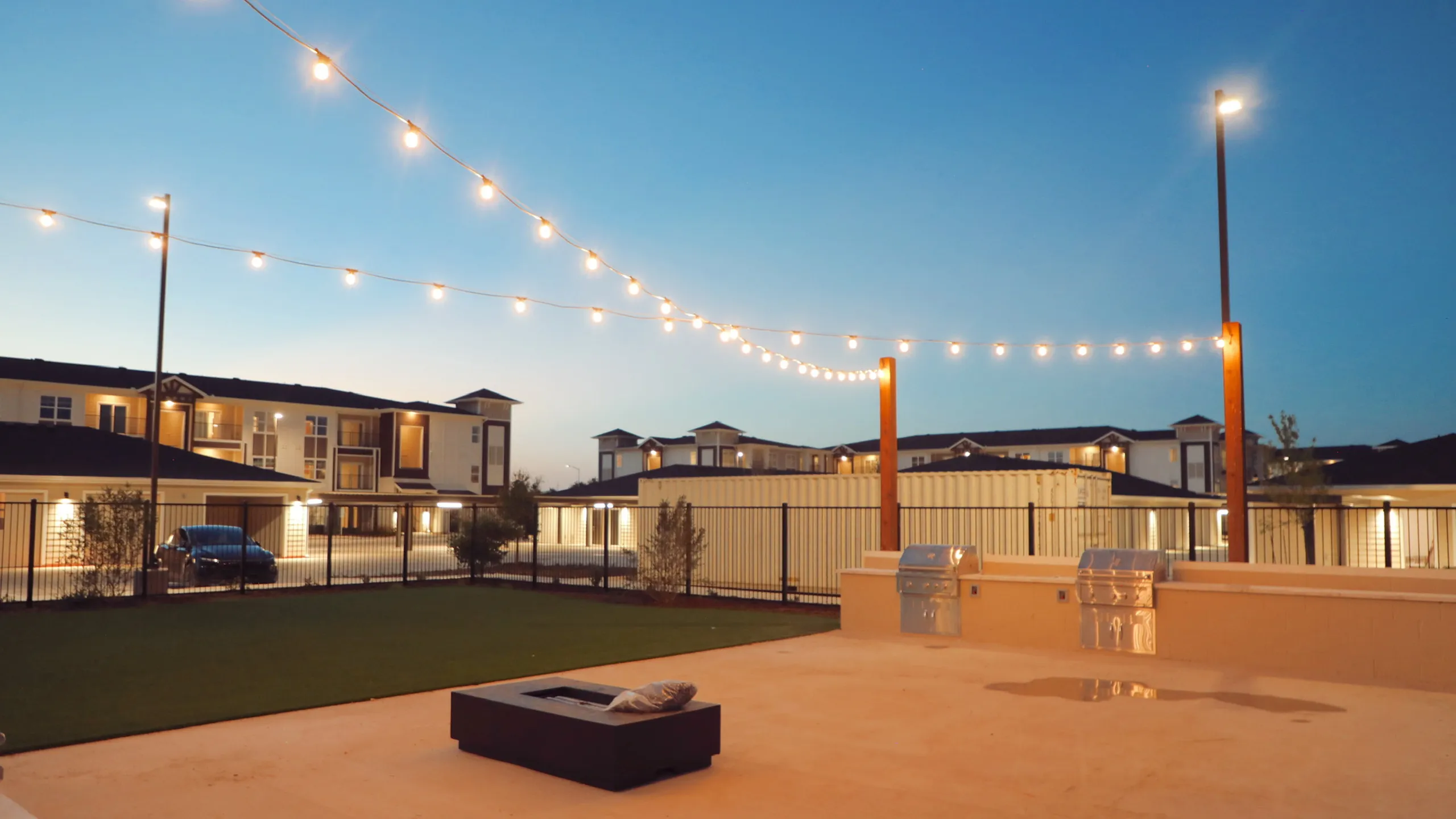 Charming outdoor space with string lights, perfect for evening gatherings, featuring a modern grill area and firepit set against stylish apartment buildings.