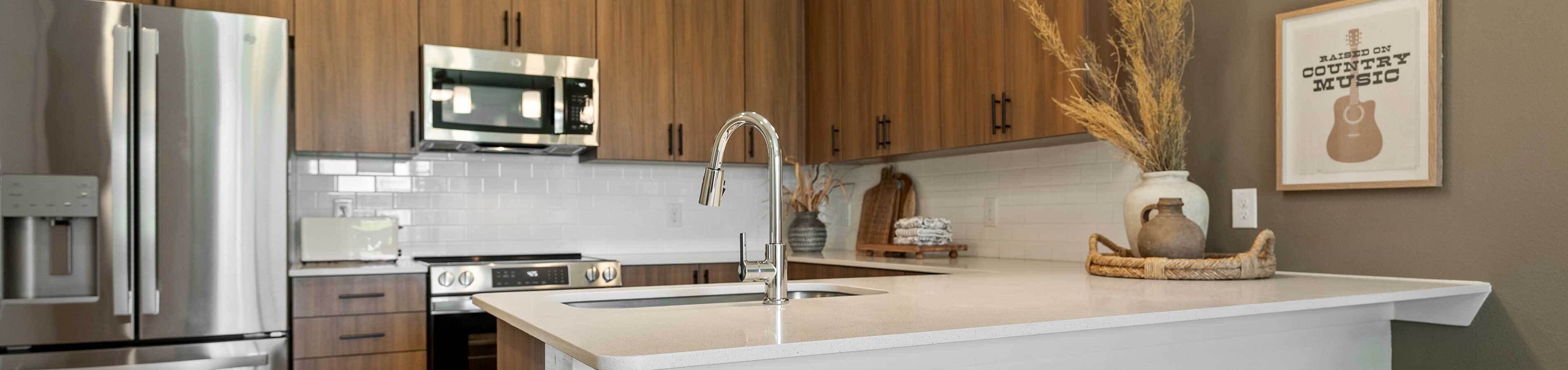 Panoramic shot of a modern luxury apartment kitchen showcasing a white quartz island countertop, chrome gooseneck faucet, warm wood-grain cabinetry, and stainless steel appliances.