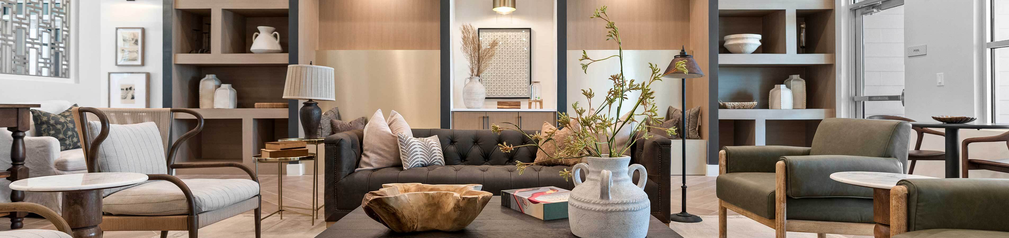 Wide-angle view of an upscale resident clubhouse lounge featuring a dark tufted chesterfield sofa, plush olive green armchairs, custom built-in wall shelving, and modern decorative accents.