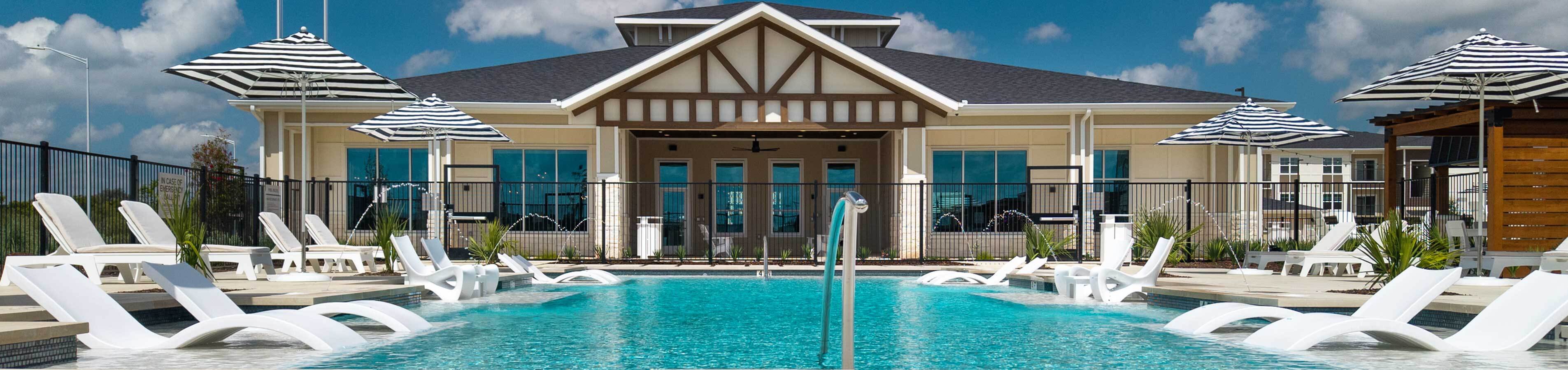 Panoramic view of a resort-style apartment swimming pool featuring submerged lounge chairs, navy-and-white striped umbrellas, and a modern community clubhouse in the background.