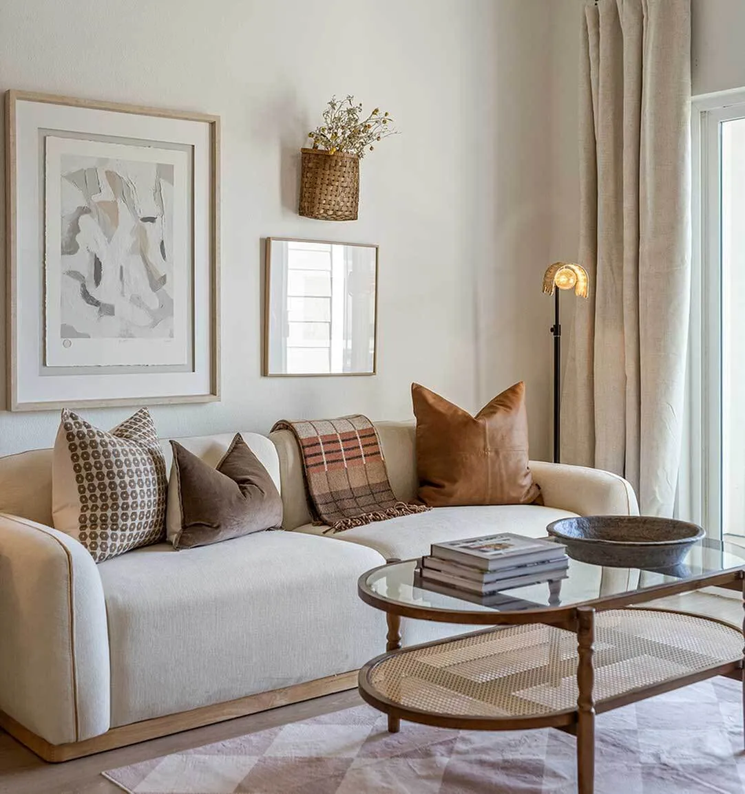 Bright, modern apartment living room featuring a cream sofa with decorative throw pillows, abstract framed wall art, and a wood-and-glass oval coffee table.