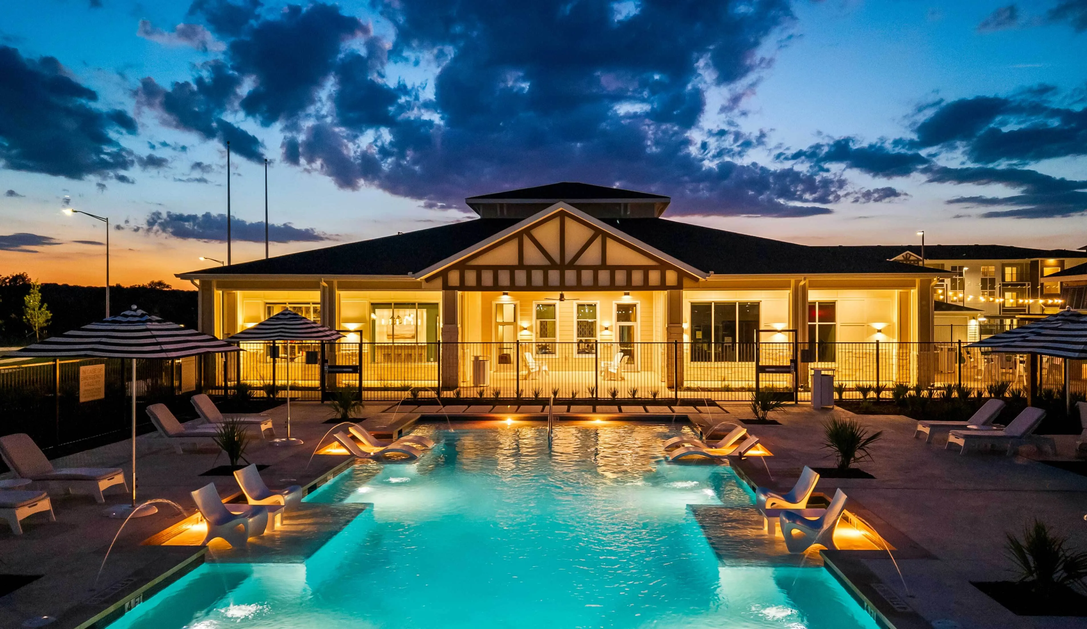 Luxurious evening poolside view at a stylish apartment complex, featuring loungers, umbrellas, and a beautifully lit building backdrop.