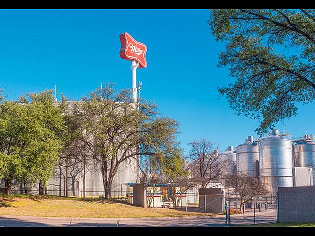 Vibrant industrial scene with large storage tanks and iconic red sign, surrounded by lush greenery and blue sky.