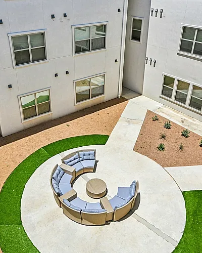 Modern apartment courtyard featuring a circular seating area with comfortable cushions, surrounded by minimalist landscaping and situated between contemporary apartments.