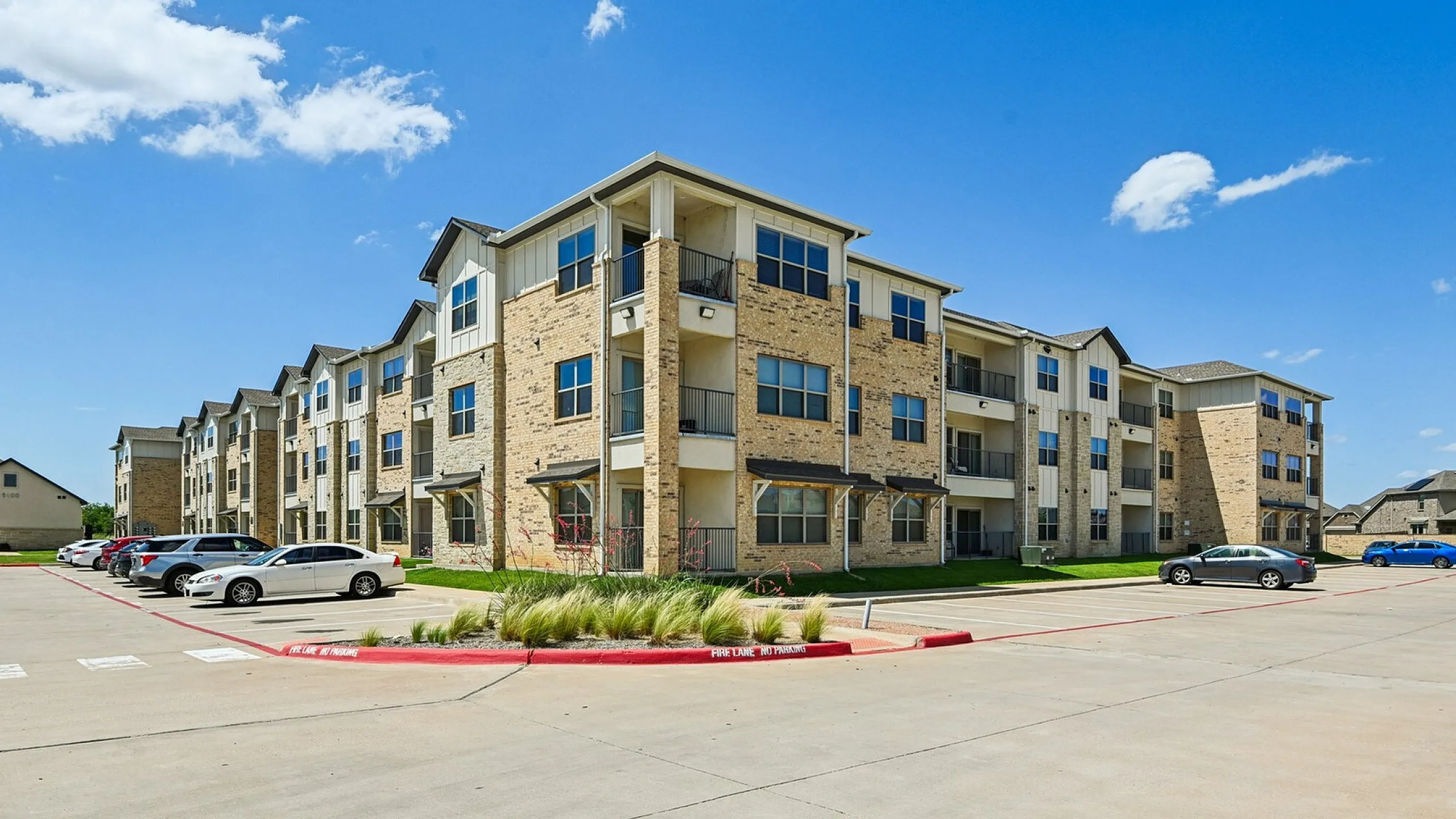 Modern three-story apartment building with brick facade, large windows, and vibrant parking area under a clear blue sky.