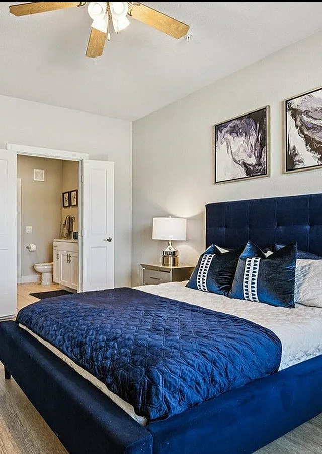 Elegant bedroom featuring a plush blue bed, decorative pillows, modern art, and view into an en-suite bathroom.